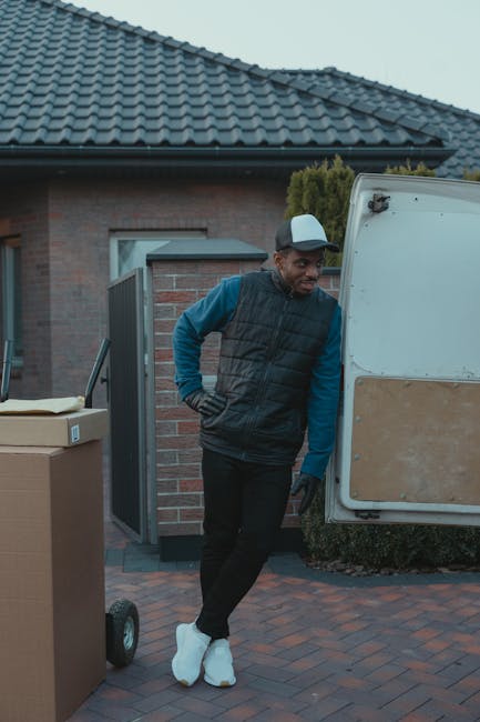 A man wearing a white cap, blue long-sleeve shirt, black quilted vest, and black trousers stands outdoors on a paved driveway next to a white moving van with a partially open rear door. He is positioned beside several packed cardboard boxes, some with packing tape, and a black hand truck is visible nearby. The scene takes place in front of a residential house with a brick exterior, a dark tiled roof, and a closed side gate. The man appears to be preparing for a home relocation, part of a furniture transport or packing and moving process, with the environment illuminated by natural daylight. This image represents the loading process during house removals carried out by Man with Van Edgware, aligned with their relocation services within the London area.