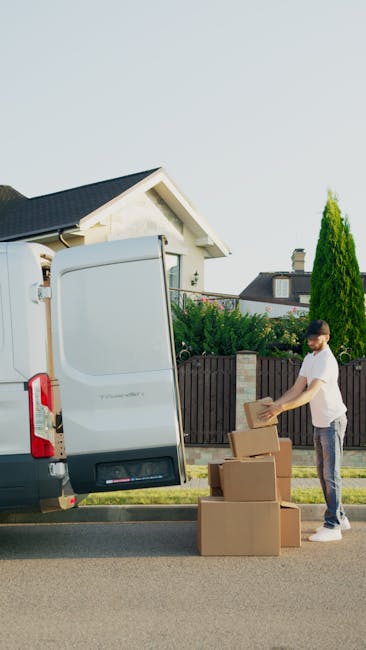 A man in casual clothing is standing on the pavement next to a stocky, white cargo van with its rear doors open, preparing for furniture transport as part of a home relocation. He is placing cardboard boxes, some wrapped in brown packing paper, onto the ground near the van. The boxes vary in size and are arranged in a small stack on the pavement. Behind the man, there is a residential property with a white facade and a dark-colored roof, along with a brown wooden fence and green shrubs. A tall, narrow evergreen tree is visible on the right side of the image, and the sky above is clear with bright natural lighting. This scene illustrates the loading process central to furniture removals and packing services offered by Man with Van Edgware, aligning with their house removals service category, focusing on efficient transport and logistics for home moves.