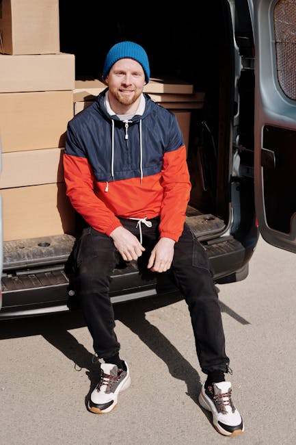 A man wearing a white cap, blue long-sleeve shirt, black quilted vest, and black trousers stands outdoors on a paved driveway next to a white moving van with a partially open rear door. He is positioned beside several packed cardboard boxes, some with packing tape, and a black hand truck is visible nearby. The scene takes place in front of a residential house with a brick exterior, a dark tiled roof, and a closed side gate. The man appears to be preparing for a home relocation, part of a furniture transport or packing and moving process, with the environment illuminated by natural daylight. This image represents the loading process during house removals carried out by Man with Van Edgware, aligned with their relocation services within the London area.