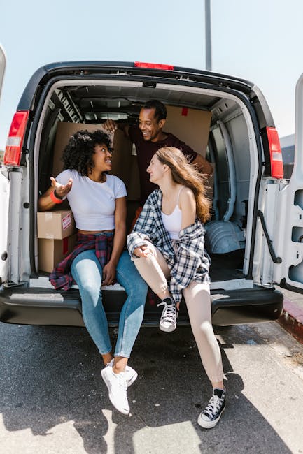 Inside the open cargo area of a large moving van parked on a driveway, two young women sit on the edge of the van's floor, engaged in conversation and smiling, while a man stands behind them, leaning into the van's interior. The women are dressed casually in T-shirts, jeans, and sneakers, with one wearing a plaid shirt tied around her waist. There are cardboard boxes and wrapped household items visible inside the cargo space, indicating a home relocation process. The van's interior is well-lit by natural light, and the exterior is painted in a neutral colour. The scene captures a moment of packing and loading during furniture transport, with the focus on the preparation for a move, consistent with house removals services offered by Man with Van Edgware, supporting the topic of moving costs and parking tips in Edgware, HA8.