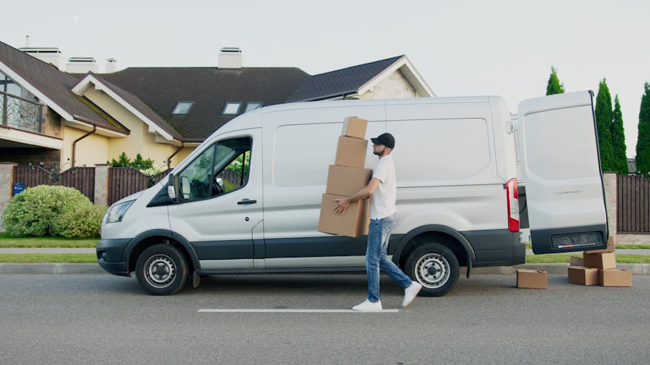 A man wearing a white t-shirt, jeans, white sneakers, and a black cap is seen loading cardboard boxes into the side door of a white cargo van parked on a residential street. The van belongs to Man with Van Edgware, a house removals service. Several additional cardboard boxes are placed on the pavement nearby, some stacked and some on the ground, ready for a home relocation or furniture transport process. The background features a modern detached house with a dark roof, large windows, a garden with neatly trimmed bushes, and a wooden fence. The scene is lit by natural daylight, indicating daytime conditions, with the van positioned parallel to the kerb, and the loading activity occurring adjacent to a well-maintained front yard.