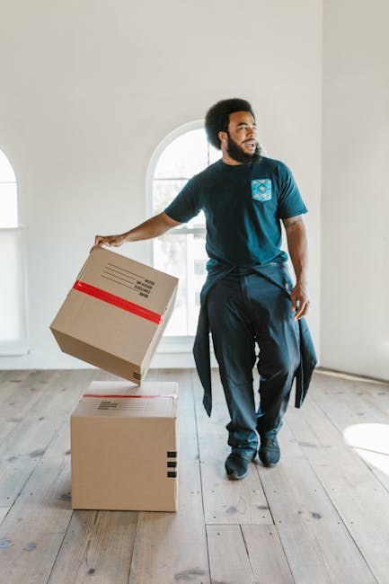 A man with dark skin, an afro hairstyle, and a beard, dressed in a dark blue t-shirt with a logo on the chest and dark trousers, is indoors on a light wooden floor near a large arched window with white frames. He is in the process of a home relocation, holding a cardboard box wrapped with red tape in his right hand, preparing to place or lift it onto or from two other stacked cardboard boxes in front of him. The environment is well-lit with natural light streaming through the window, revealing a minimalistic room with white walls and a clean, uncluttered space. This scene illustrates the packing and moving process with attention to furniture and packing materials, fitting within the services offered by Man with Van Edgware, specialists in removals and furniture transport for small house moves. The image emphasizes careful handling of boxes as part of a professional home relocation or moving logistics service.
